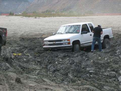 1987 Ford Bronco - A white pickup truck is stuck deep in the mud, with a person attempting to assess the situation.