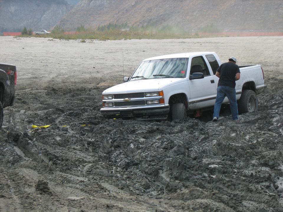 White pickup truck stuck in mud with a person standing next to it. Another truck is partially visible on the left