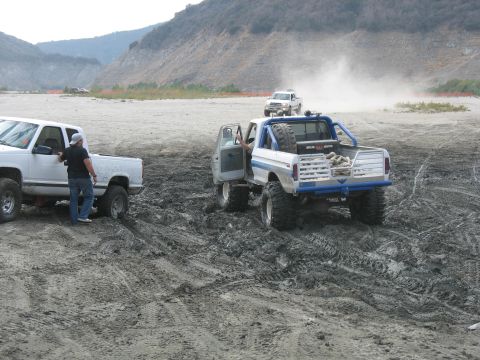1987 Ford Bronco - Two trucks navigate a muddy terrain with hills in the background.