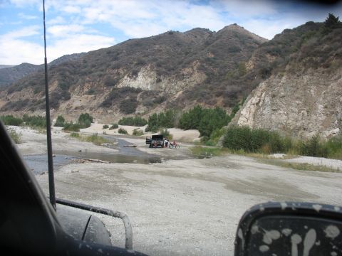 1987 Ford Bronco - A truck and motorcycle are seen in a dry riverbed with mountains in the background.