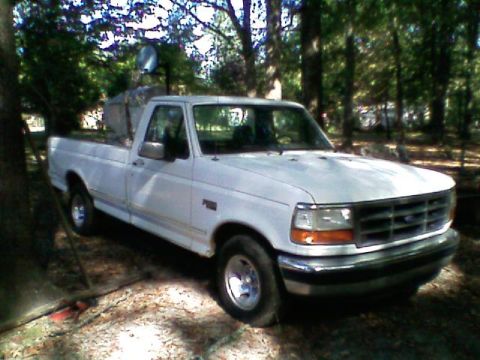 1992 Ford F-150 - White Ford F-Series regular-cab long-bed pickup parked in a shaded wooded yard, shown from the passenger-side front three-quarter angle. It…