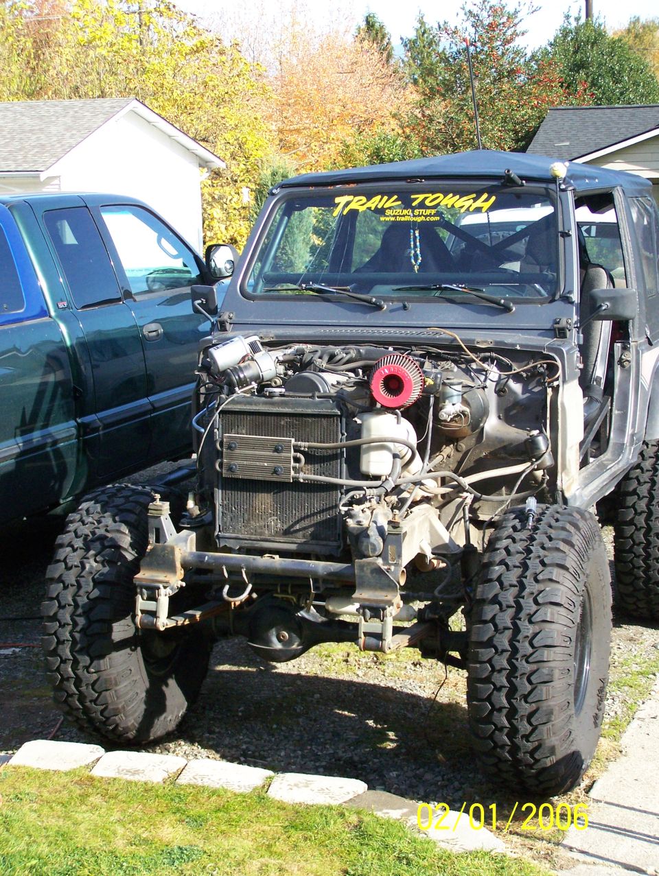 Gray 1988 Suzuki Samurai, front view, engine exposed, off-road tires, parked next to a green pickup truck