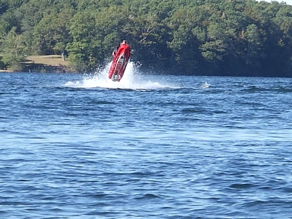 Red personal watercraft nearly vertical in white spray on a lake with wooded shoreline behind