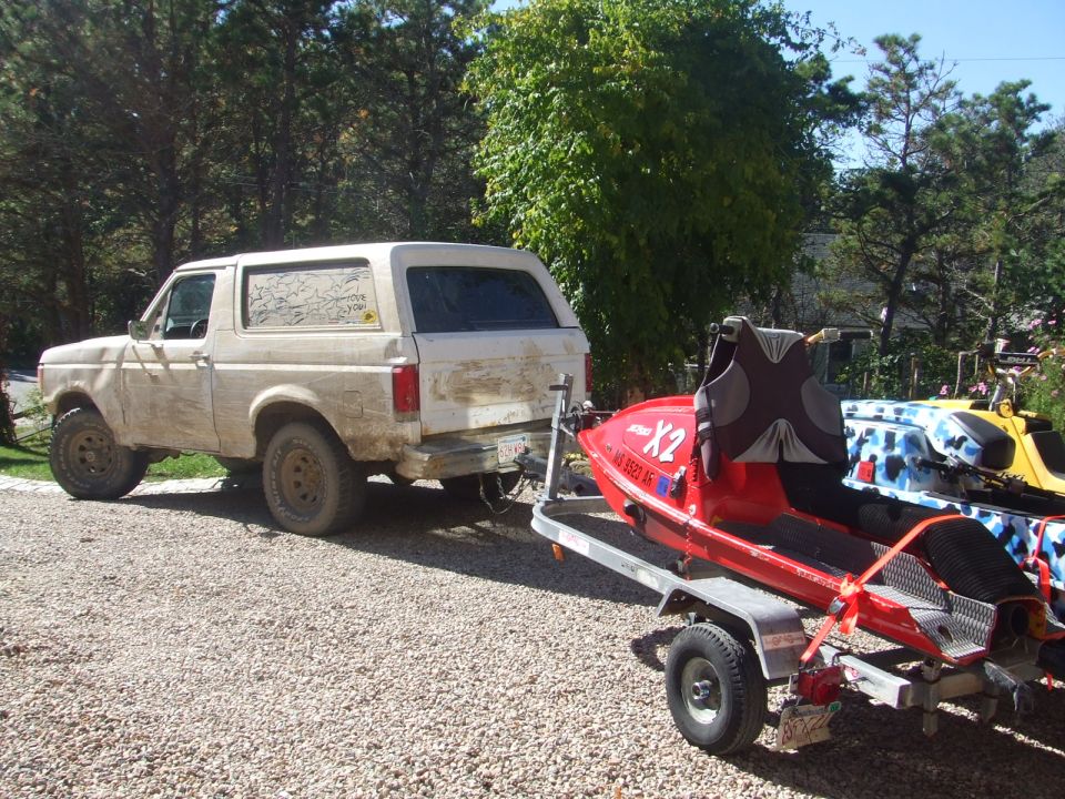 Red stand-up jet ski on a trailer behind a muddy white Ford Bronco on a gravel lot