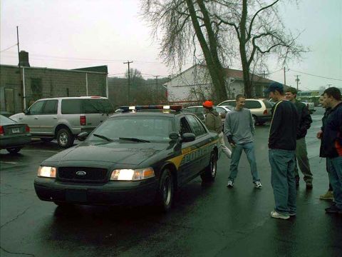 2000 Ford Crown Victoria - Ford Crown Victoria police cruiser with roof light bar in a parking lot, people standing alongside it.