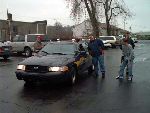 2000 Ford Crown Victoria - A black 2000 Ford Crown Victoria police car is parked in a lot with several people standing around it.