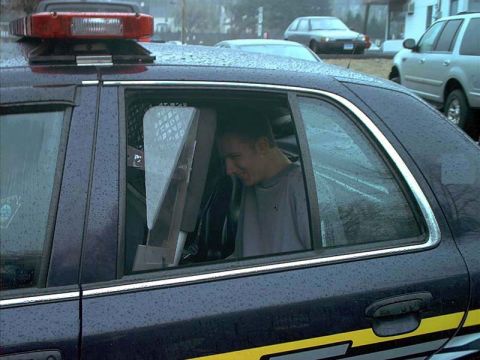 2000 Ford Crown Victoria - Close side view of a rain-beaded dark blue Ford Crown Victoria police car with a red roof lightbar and yellow body stripe, showing a person…