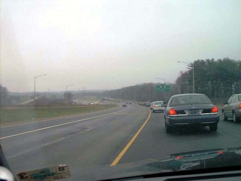 2000 Ford Crown Victoria - Windshield view from inside a car following a dark blue Ford Crown Victoria on a wet divided highway, with brake lights lit and a green ove…