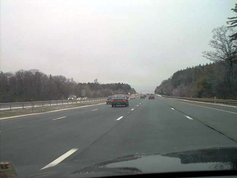 Cars driving on a highway under an overcast sky. Trees line the right side of the road
