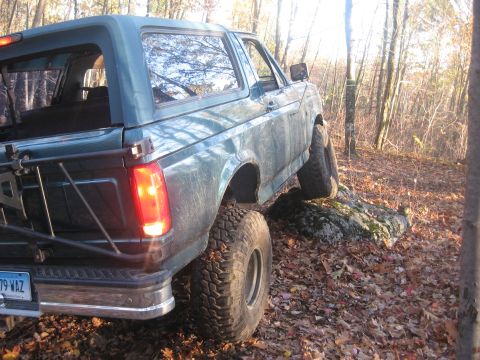 1996 Ford Bronco - A teal Ford Bronco navigates rocky terrain in a wooded area, its off-road tires gripping the uneven surface.