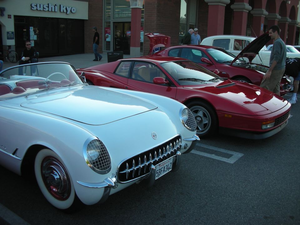 White vintage Corvette beside a red Ferrari Testarossa at an outdoor car meet