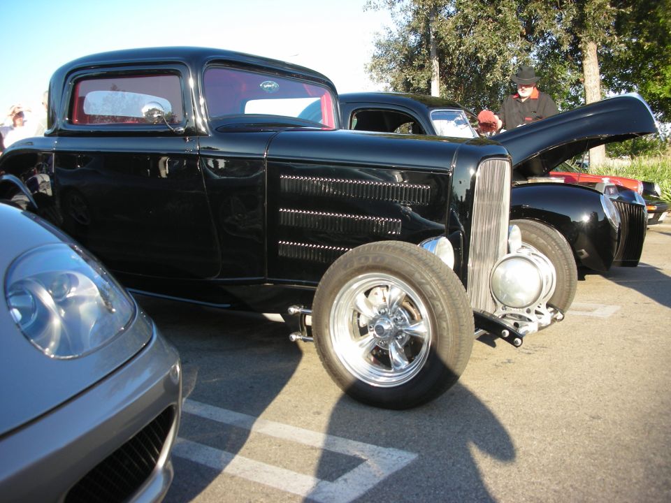 Black hot rod coupe in profile with chrome five-spoke wheels at a sunny car meet
