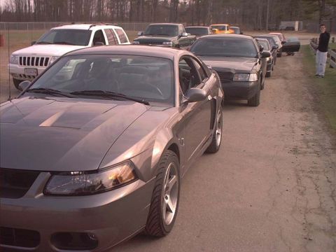 2000 Ford Crown Victoria - A line of cars, including a Ford Mustang, are parked on a dirt road. A person stands to the side.