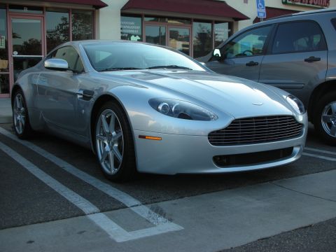 1998 Ford Crown Victoria - silver Aston Martin V8 Vantage coupe parked front three-quarter outside a storefront with reflective glass.