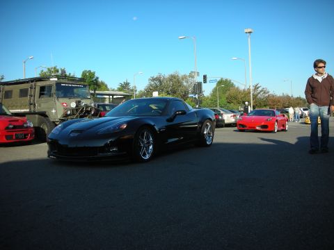 1998 Ford Crown Victoria - black C6 Corvette coupe parked in a lot, with a red Ferrari, a man, and an olive military truck in the background.