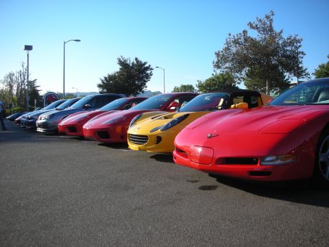 1998 Ford Crown Victoria - lined-up row of sports cars at a morning meet, led by a red Corvette with a yellow car and others stretching back.
