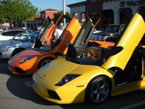 1998 Ford Crown Victoria - orange and a yellow Lamborghini Murcielago sit side by side with scissor doors raised at a car meet outside shops.