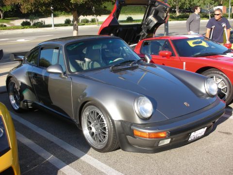 1998 Ford Crown Victoria - gunmetal-gray Porsche 911 with wide flared arches and mesh wheels sits parked beside a red 911 with its hood open.