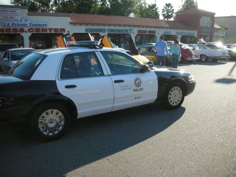 1998 Ford Crown Victoria - black-and-white LAPD Crown Victoria police car sits in a street lined with shops and parked classic cars.