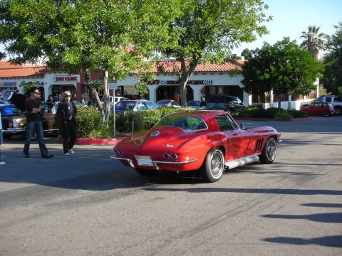 1998 Ford Crown Victoria - red split-era Corvette Sting Ray coupe rolls through a parking lot past onlookers and shops on a sunny day.