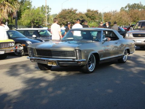 1998 Ford Crown Victoria - silver-gold 1960s Buick Riviera coupe with stacked headlights and whitewall tires sits in a crowded car-meet lot.