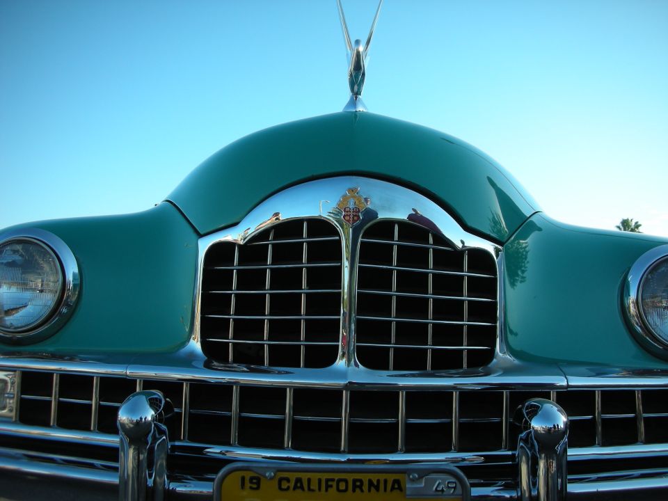 Close-up of a green vintage car's chrome grille and hood ornament against blue sky