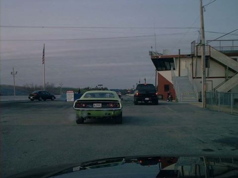2000 Ford Crown Victoria - A green muscle car is seen from the rear at a drag strip, with a black SUV in the background.