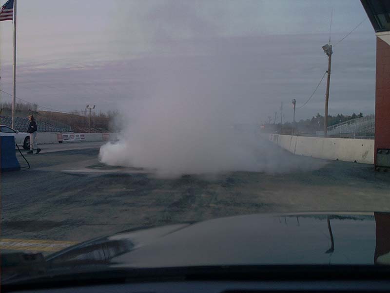 Car doing a burnout on a drag strip, emitting a large cloud of smoke. A person stands near a white car in the background