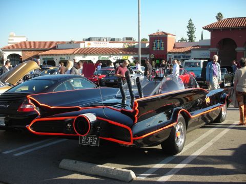1998 Ford Crown Victoria - black Batmobile replica with red trim and a chrome exhaust nozzle parked among other cars at a strip-mall car gathering.