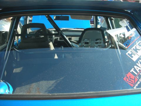 1998 Ford Crown Victoria - View through the rear glass of a blue car revealing a roll cage, racing bucket seats, and sponsor stickers on the windows.