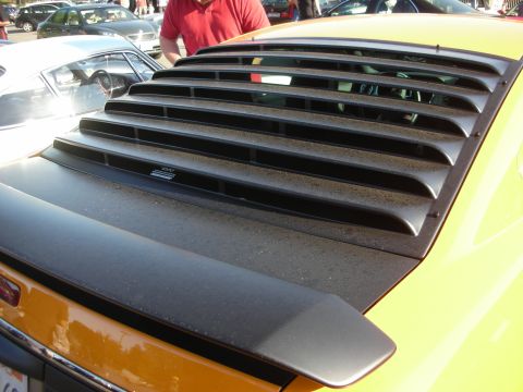 1998 Ford Crown Victoria - Detail of black rear-window louvers and rear spoiler mounted on an orange-yellow car at an outdoor gathering.