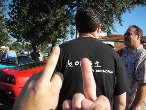 1998 Ford Crown Victoria - man seen from behind wearing a black T-shirt reading 'boost - my anti-drug' as two hands point at him; red sports car nearby.