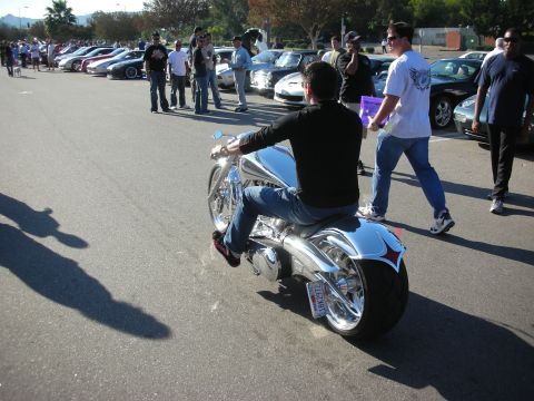 1998 Ford Crown Victoria - man rides a chrome custom chopper motorcycle with a fat rear tire through a parking lot full of cars and onlookers.