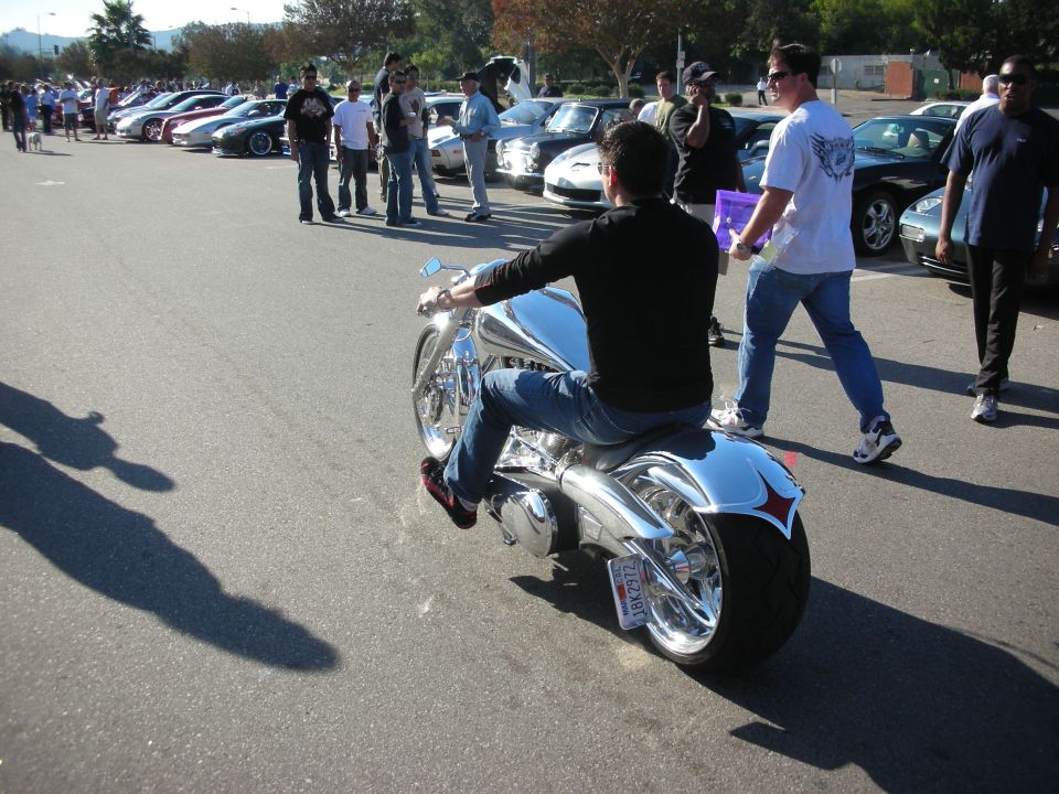 Man riding a chrome chopper motorcycle through a crowded car-show lot