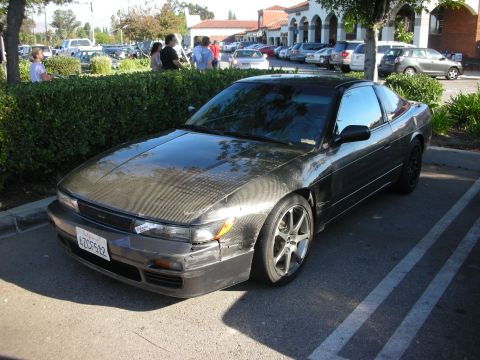 1998 Ford Crown Victoria - weathered black Nissan coupe with a carbon-fiber hood and gray wheels parked along a hedge in a sunny lot.