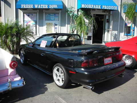 1998 Ford Crown Victoria - A black convertible Mustang is parked in front of a store with a 'For Sale' sign in the window.