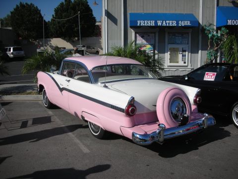 1998 Ford Crown Victoria - A classic pink and white Ford Fairlane coupe is parked next to a black convertible with a 'For Sale' sign.