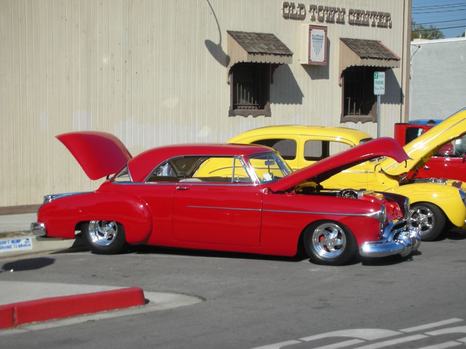 Red lowered custom coupe with open hood parked beside a yellow coupe in front of Old Town Center building