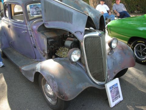 1998 Ford Crown Victoria - A custom hot rod, possibly a 1934 Ford, is displayed with its hood open, revealing the engine. A sign in front provides more details.