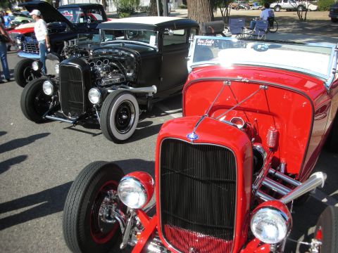 1998 Ford Crown Victoria - Two classic hot rod cars are parked side-by-side at a show, with a 'For Sale' sign visible on the red car's windshield.