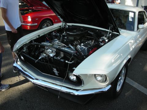 1998 Ford Crown Victoria - A white 1969 Ford Mustang is displayed with its hood open, revealing a detailed engine bay. Another red classic car is visible in the backg…