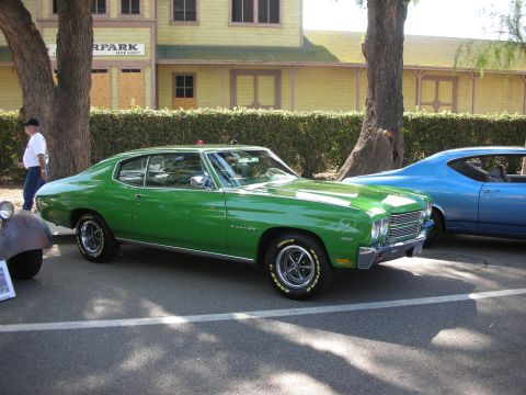 1998 Ford Crown Victoria - A green 1970 Chevrolet Chevelle Malibu is parked next to a blue car in front of a yellow building.