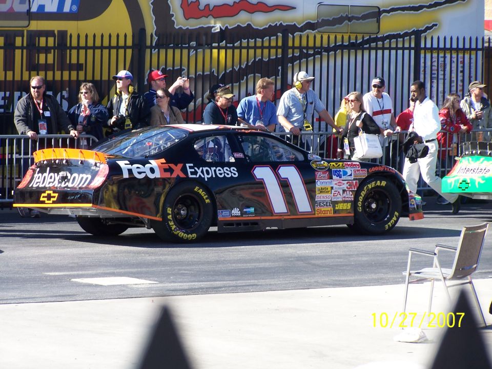 Black FedEx Express race car with number 11 on its side, parked behind a fence with spectators