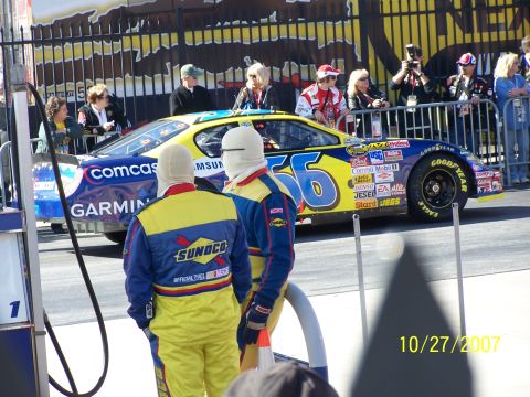 1979 Ford F-150 - A NASCAR race car with sponsors like Comcast and Garmin is seen from the side, with two crew members in yellow and blue suits standing near…