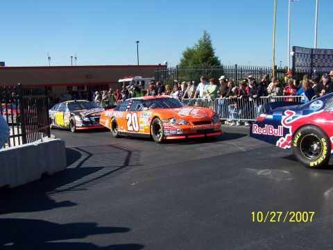 1979 Ford F-150 - Multiple NASCAR stock cars are lined up on a track with spectators watching from behind a fence.