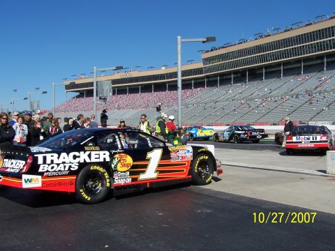 1979 Ford F-150 - Several NASCAR stock cars are staged on the pit lane with spectators in the grandstands under a clear blue sky.