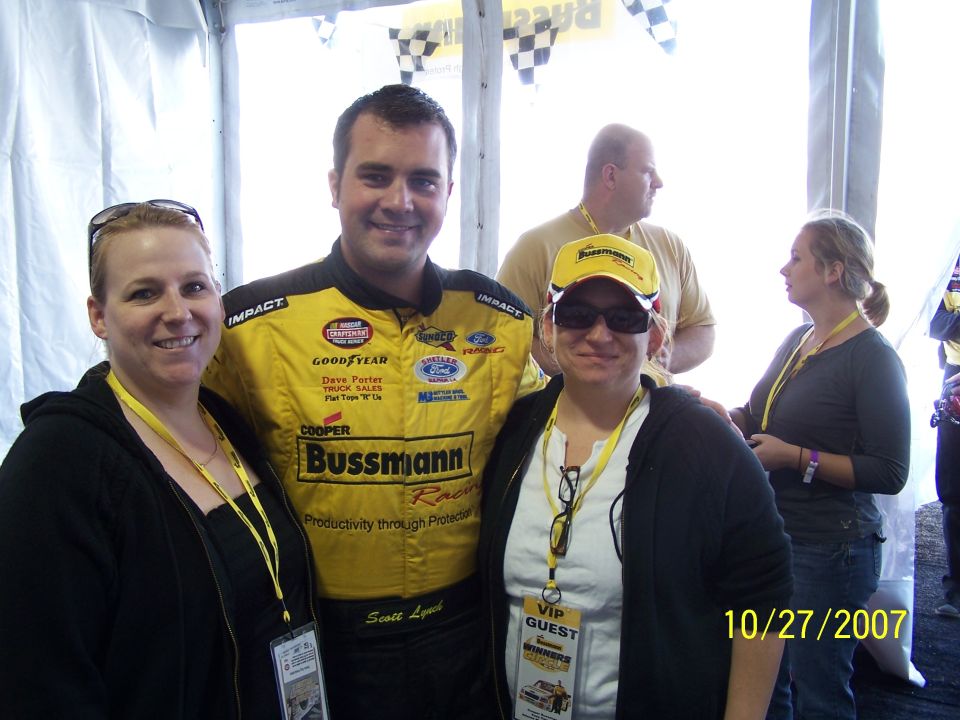 Three people posing for a photo at a race event. One person wears a yellow racing suit with sponsor logos