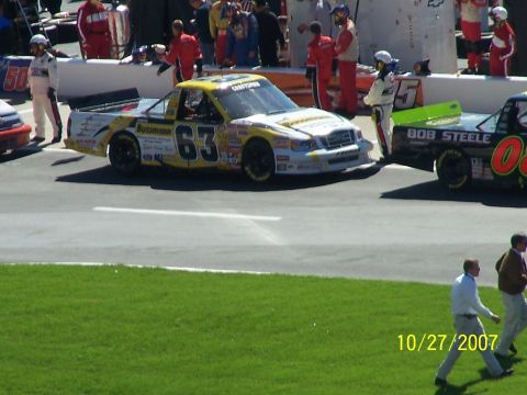 1979 Ford F-150 - Race trucks line up on pit road with crew members and officials nearby.