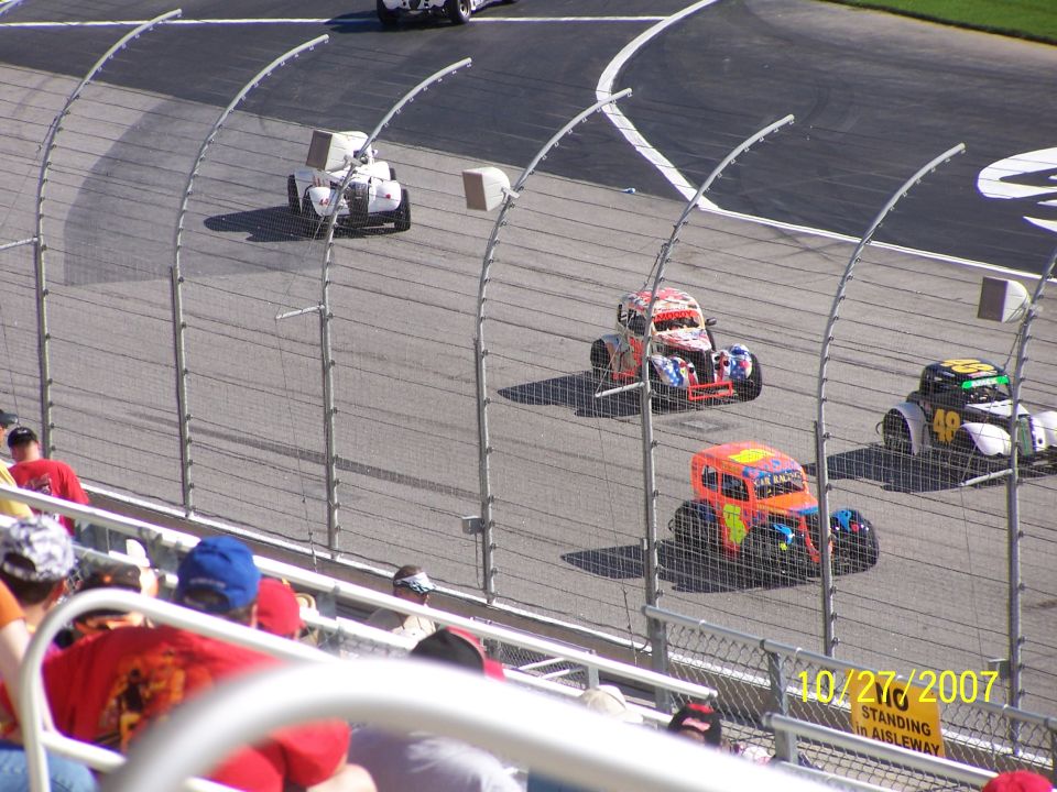 Stock cars racing on a track, one with orange and blue livery, another with red and white