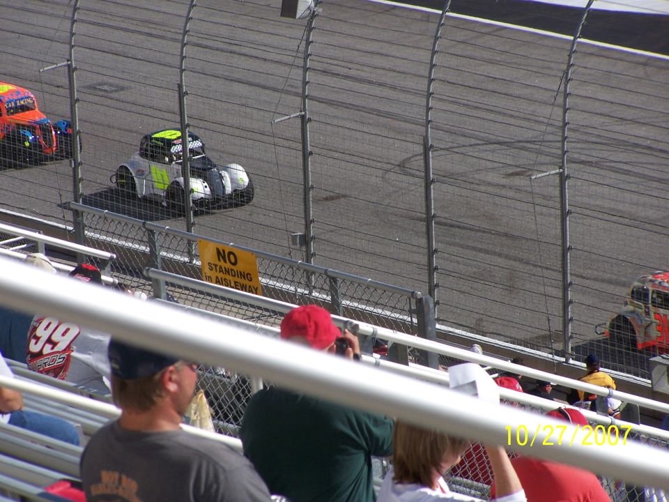 Stock cars racing on a track, viewed from the stands. A sign reads 'NO STANDING IN AISLEWAY'